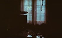 A dimly lit room with sheer curtains covering a window that lets in diffused natural light. The outlines of objects such as a table and a monitor or television are faintly visible. The overall ambiance is dark with a soft, muted glow coming through the window.