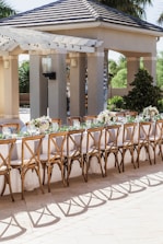 An outdoor seating arrangement with a long table set for an event. The table is elegantly decorated with white tablecloths, floral centerpieces, and wine glasses. Wooden chairs with intricate designs surround the table. The background includes a pavilion structure with columns and a tiled roof, surrounded by greenery.