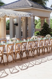 An outdoor seating arrangement with a long table set for an event. The table is elegantly decorated with white tablecloths, floral centerpieces, and wine glasses. Wooden chairs with intricate designs surround the table. The background includes a pavilion structure with columns and a tiled roof, surrounded by greenery.