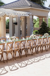 An outdoor seating arrangement with a long table set for an event. The table is elegantly decorated with white tablecloths, floral centerpieces, and wine glasses. Wooden chairs with intricate designs surround the table. The background includes a pavilion structure with columns and a tiled roof, surrounded by greenery.