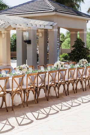 An outdoor seating arrangement with a long table set for an event. The table is elegantly decorated with white tablecloths, floral centerpieces, and wine glasses. Wooden chairs with intricate designs surround the table. The background includes a pavilion structure with columns and a tiled roof, surrounded by greenery.