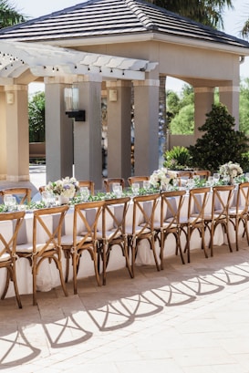 An outdoor seating arrangement with a long table set for an event. The table is elegantly decorated with white tablecloths, floral centerpieces, and wine glasses. Wooden chairs with intricate designs surround the table. The background includes a pavilion structure with columns and a tiled roof, surrounded by greenery.