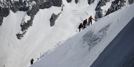 A group of climbers is ascending a steep, snow-covered mountain slope. The scene features rugged terrain with jagged rocks and deep snow, and the climbers are equipped with helmets and gear, indicating a high-altitude trek. The backdrop is mountainous with sharp ridges and white snow, suggesting a remote and challenging environment.
