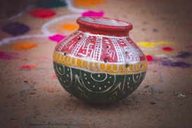 A traditional clay pot is intricately painted with colorful designs featuring red, white, yellow, and green. The pot is placed on a sandy surface, surrounded by vibrant rangoli patterns in the background, featuring various bright colors like purple, pink, and orange.