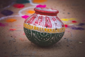 A traditional clay pot is intricately painted with colorful designs featuring red, white, yellow, and green. The pot is placed on a sandy surface, surrounded by vibrant rangoli patterns in the background, featuring various bright colors like purple, pink, and orange.
