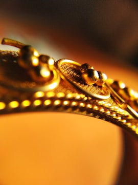 A close-up view of a gold object, possibly jewelry, featuring intricate detailing with small spherical elements and textured patterns.