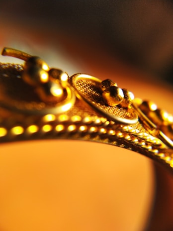 A close-up view of a gold object, possibly jewelry, featuring intricate detailing with small spherical elements and textured patterns.