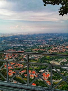 The image showcases a panoramic view from a high vantage point, overlooking a sprawling landscape with a mix of urban and rural elements. There are clusters of buildings with red roofs scattered across a green terrain. The background features rolling hills and a horizon with expansive skies, partially covered by clouds. A metal railing in the foreground adds depth to the scene.