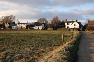 A rural landscape with several traditional English cottages surrounded by fields. The scene is bathed in warm sunlight, with a clear sky and some scattered clouds above. A narrow country road leads up to the houses, which are lined by a wooden fence. Trees without leaves can be seen in the background, suggesting it might be early spring or late autumn.