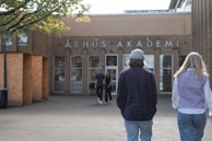 Two people are walking towards a brick building with the sign 'ÅRHUS AKADEMI' at the entrance. The scene is set in daylight, and the entrance is framed by trees with green leaves on the left.