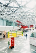 A spacious indoor area with a modern, geometric ceiling featuring metal trusses. In the foreground, there is an illuminated signpost with various directions and labels for a restaurant, café, and bar. Above, a vintage red biplane is suspended from the ceiling, adding an element of historical interest. People are walking and standing around, with one person in the foreground checking their phone.