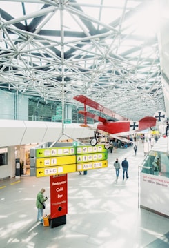 A spacious indoor area with a modern, geometric ceiling featuring metal trusses. In the foreground, there is an illuminated signpost with various directions and labels for a restaurant, café, and bar. Above, a vintage red biplane is suspended from the ceiling, adding an element of historical interest. People are walking and standing around, with one person in the foreground checking their phone.