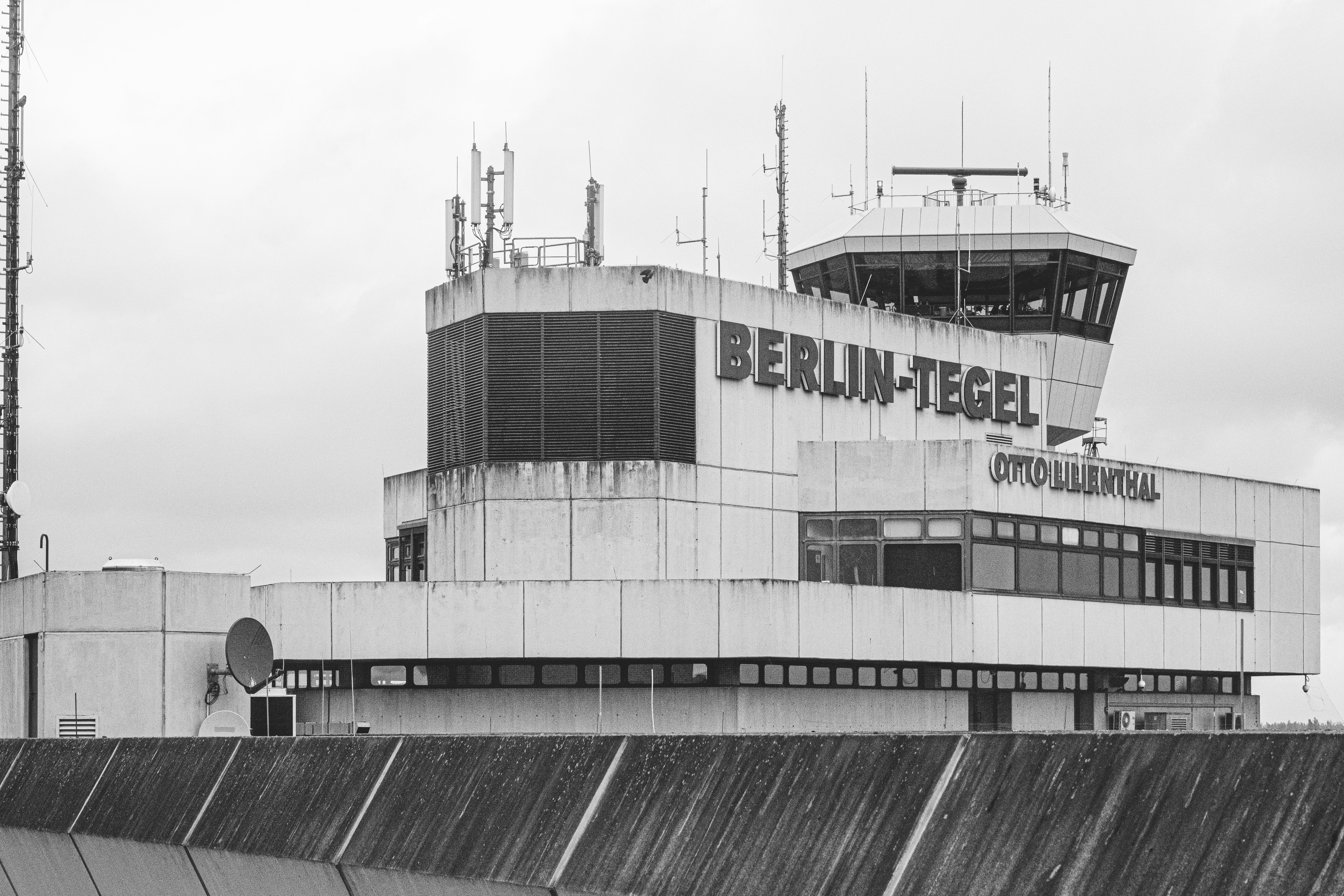 Monochrome photograph of Berlin-Tegel’s control tower rising above the terminal, with rain-slicked surfaces and bold signage. The composition emphasizes industrial geometry against a moody sky.