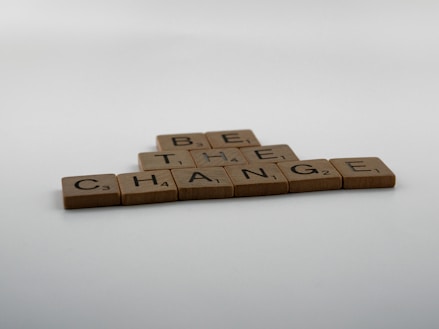 Scrabble tiles arranged on a flat surface spelling out the phrase 'Be the Change' in a pyramid shape. The background is a plain, off-white color with a soft gradient.