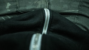 A close-up view of a black fabric with a silver zipper running down the middle, surrounded by dark camouflage-patterned pants.