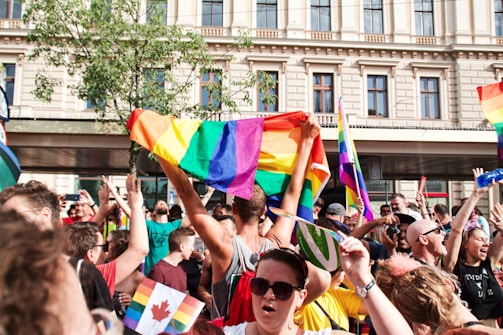 A large group of people is celebrating outdoors, many of them holding rainbow flags. They are gathered in front of a building with ornate architecture, and there are trees in the background. Many participants are smiling and appear to be enjoying the event.