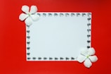 A white rectangular card is adorned with a border of small heart-shaped gems. Two white flowers are positioned at the top left and bottom right corners, providing a delicate touch against a vibrant red background.