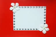 A white rectangular card is adorned with a border of small heart-shaped gems. Two white flowers are positioned at the top left and bottom right corners, providing a delicate touch against a vibrant red background.