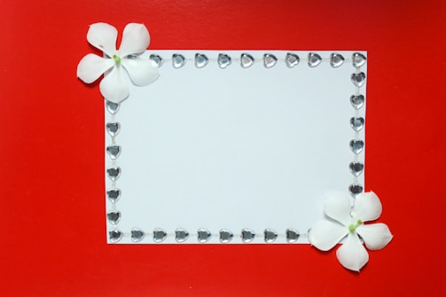 A white rectangular card is adorned with a border of small heart-shaped gems. Two white flowers are positioned at the top left and bottom right corners, providing a delicate touch against a vibrant red background.