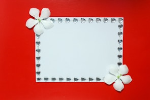 A white rectangular card is adorned with a border of small heart-shaped gems. Two white flowers are positioned at the top left and bottom right corners, providing a delicate touch against a vibrant red background.