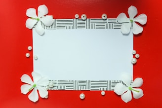 A rectangular white card bordered by a patterned black and white design is surrounded by white flowers and scattered pearls on a vibrant red background.
