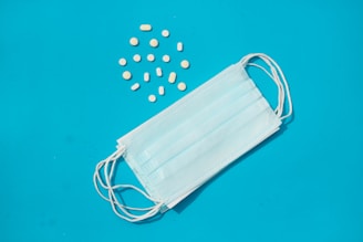 A light blue surgical mask is placed on a bright blue background with several small, white and yellow pills scattered nearby.