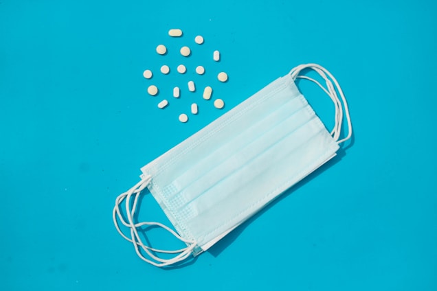 A light blue surgical mask is placed on a bright blue background with several small, white and yellow pills scattered nearby.