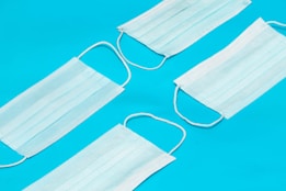Four disposable face masks are arranged on a bright blue background. The masks are white with elastic ear loops and are positioned in a diagonal layout.
