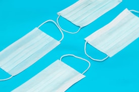 Four disposable face masks are arranged on a bright blue background. The masks are white with elastic ear loops and are positioned in a diagonal layout.