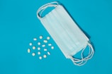 A blue medical mask lies on a blue background next to scattered yellow and white pills in various shapes and sizes.
