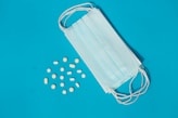 A blue medical mask lies on a blue background next to scattered yellow and white pills in various shapes and sizes.