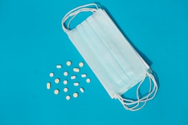 A blue medical mask lies on a blue background next to scattered yellow and white pills in various shapes and sizes.