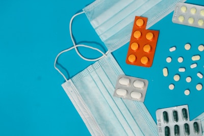 A collection of medical items including two blue surgical masks and several blister packs of pills arranged on a bright blue surface. Orange, green, and white pills are scattered around the blister packs.