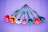 A vibrant arrangement of seven paper flowers, each with green stems and leaves. The flowers feature striped petals in a variety of bright colors including red, orange, pink, white, blue, and purple. They are laid out against a rich purple textured background.