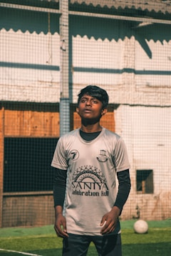 A person is standing on a grassy field, wearing a sports jersey with logos and text on it. The individual looks focused, possibly watching something above them. The background includes a net and a partially visible building with a wooden door.