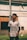 A person is standing on a grassy field, wearing a sports jersey with logos and text on it. The individual looks focused, possibly watching something above them. The background includes a net and a partially visible building with a wooden door.