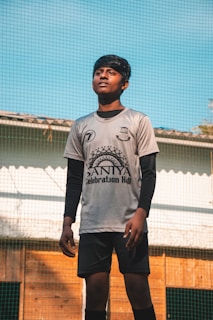A young person in a sports jersey is standing outdoors. They appear to be on a field with a net in the background. The person is looking slightly upwards with a focused expression.