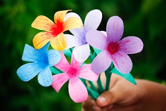 A hand holds a colorful bouquet of paper flowers against a blurred green background. The flowers come in a variety of colors including pink, blue, yellow, and white with red centers. Each flower has five petals and is attached to green stems.