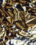 A close-up view of crumpled or wavy metallic foil, with a reflective golden and silver surface creating abstract patterns and highlights.