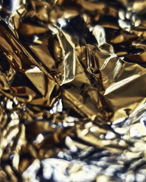 A close-up view of crumpled or wavy metallic foil, with a reflective golden and silver surface creating abstract patterns and highlights.