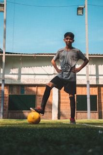 A young person is standing on a soccer field with one foot resting on a yellow soccer ball. They are wearing a gray soccer jersey and black shorts with knee-high socks. The background features a net and a wooden wall with a blue sky overhead.