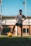 A young person is standing on a soccer field with one foot resting on a yellow soccer ball. They are wearing a gray soccer jersey and black shorts with knee-high socks. The background features a net and a wooden wall with a blue sky overhead.
