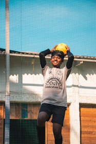 A young person wearing a sports uniform holds a yellow soccer ball above their head, standing on a sports field with netting visible in the background. The lighting is bright, suggesting a sunny day.