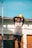 A young person wearing a sports uniform holds a yellow soccer ball above their head, standing on a sports field with netting visible in the background. The lighting is bright, suggesting a sunny day.
