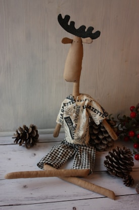A handmade fabric moose toy with a long nose and black antlers sits among pinecones. It is wearing a shirt with newspaper print patterns and checkered shorts. The scene is set against a wooden background, and some red berries are visible in the corner.