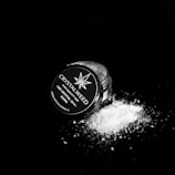 A small jar labeled 'CRYSTALWEED' lies on its side against a dark background, with a fine white powder spilling out onto the surface. The label indicates the product is a high-quality CBD crystal.