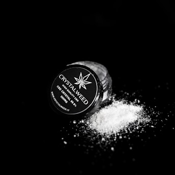 A small jar labeled 'CRYSTALWEED' lies on its side against a dark background, with a fine white powder spilling out onto the surface. The label indicates the product is a high-quality CBD crystal.
