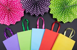 A collection of colorful paper shopping bags arranged in a row on a black background. Above them are large, polka-dotted paper fan decorations in vivid colors like pink, purple, and green.