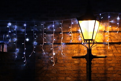 A brick wall is adorned with strings of white fairy lights. A lamp post casts a warm yellow glow against the wall, creating a contrasting blend of cool and warm light.
