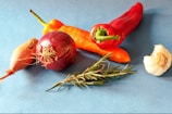 An arrangement of fresh vegetables and herbs on a blue background, including a red onion, garlic bulb, red and orange peppers, and a sprig of rosemary.